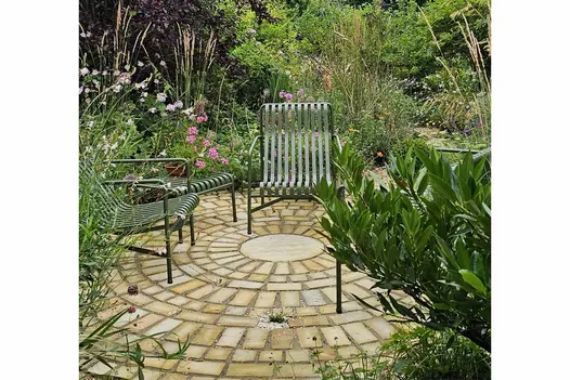 Four green metal armchairs arranged in a circle rest on Mint Sandstone Setts, surrounded by trees, shrubs, and lush greenery.