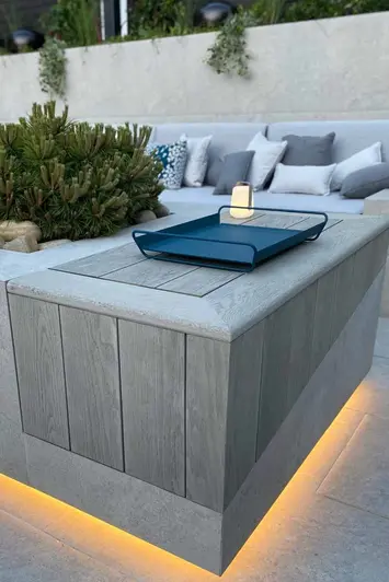 Close up of raised wall clad with smoked oak millboard with lighting underneath and drinks tray on top, built in sofa in distance.