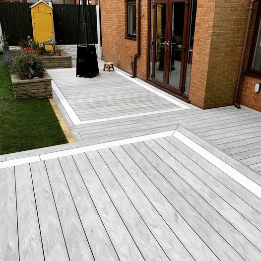 Red brick house leads to decking area of luna designboard with raised brick flowerbeds and small yellow shed in the background.