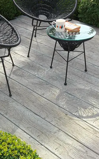 2 black bucket chairs and table cast shadows on Driftwood Millboard decking edged with low trimmed hedge.