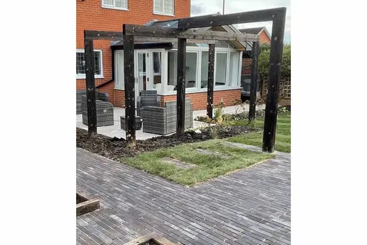 Back garden of modern brick house, laid to lawn and Ancona Dutch clay pavers with wood pergola over beds next to patio.