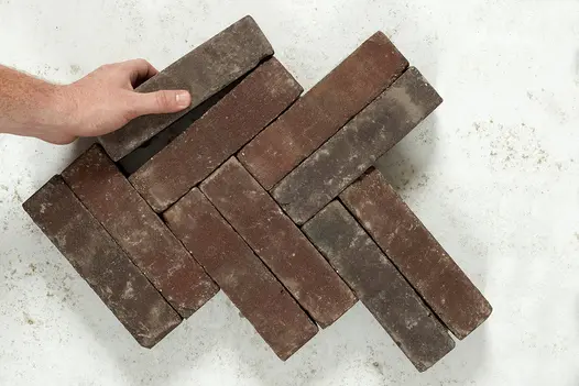 Hand places single dark brown Bergamo clay paver against 9 laid herringbone on light background. Free UK delivery available.