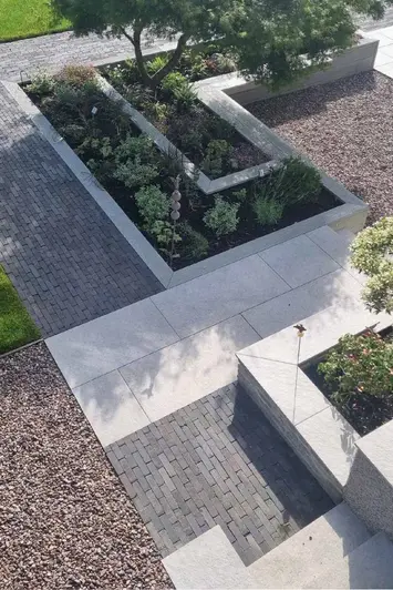 Clean cut porcelain slabs surrounded by pebbles, with chunks of Lugano Dutch Clay Pavers between flowerbeds.