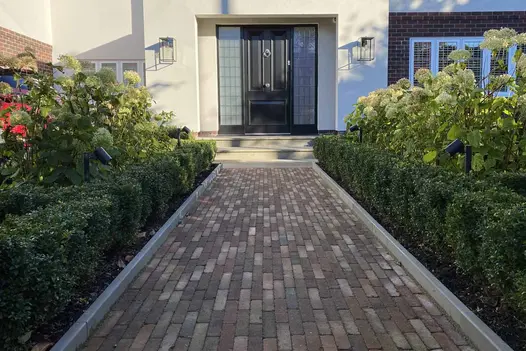 View from shadows show heath edging stones run adjacent to hedges either side of clay paver path leading up to front door.