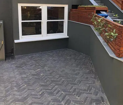 Small corner patio of amersham clay pavers in herringbone pattern sits against dark wall with red bricks on top.