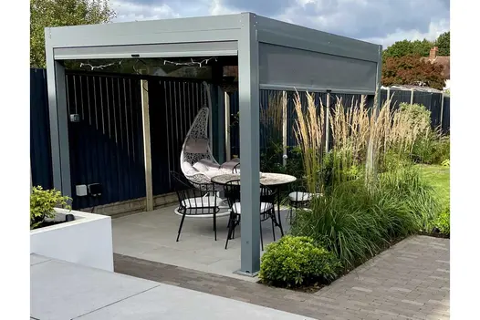 Pergola-covered patio of Florence Grey porcelain paving. Lawn and beds beyond. Design by Emily’s Garden. Built by JRLandscaping.