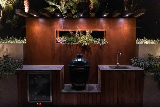 Night view of outdoor kitchen backed by high wall faced with Corten Steel external cladding with cubby hole holding plants.