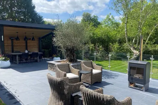 Rattan armchairs overlook lawn on wide Florence Dark porcelain patio with glass balustrade. Design by Thames Valley Landscapes.