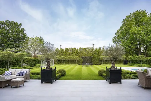Patio of Florence Grey porcelain paving gives access to lawn between twin square planters with olive trees. Swimming pool on right.
