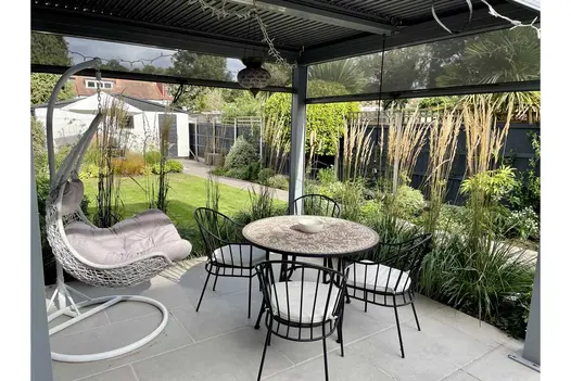 Dining set on Florence Grey Porcelain paving beneath Dark Grey Metal pergola, edged by lawn, beds and clay pavers. 