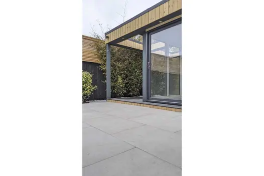 Glass-fronted garden office surrounded by Florence grey porcelain paving. Tall fence topped with slatted wood panels in background.