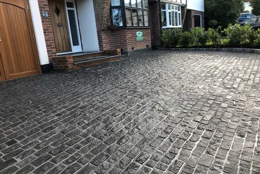 Natural Black Granite setts in wet weather used on driveway, leading up to the front of a house, bushes in the background.