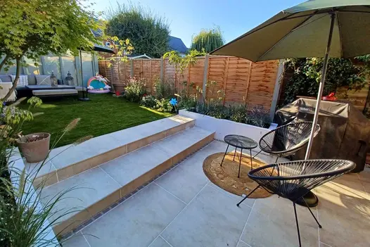 By fence, 2 wide Cream porcelain steps with 5mm chamfer edge rise from matching paving to lawn. Built by Kingston Landscape Group.
