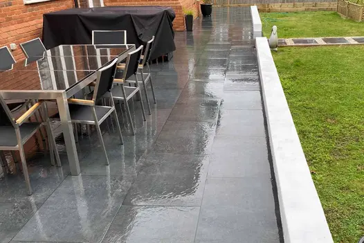 Black and silver garden dining set sits on Charcoal porcelain paving slab patio with low white wall creating border with lawn. 