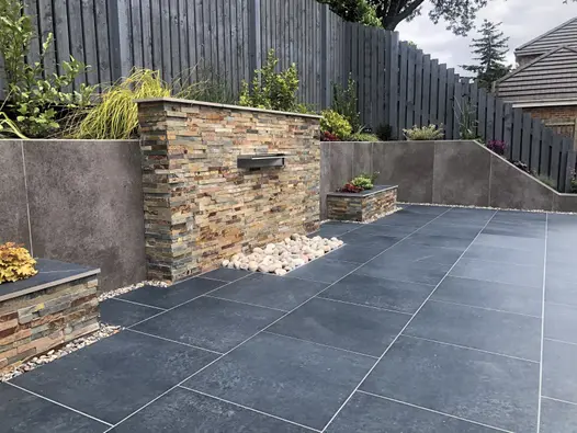 Charcoal porcelain paving patio in front of a stone clad water feature and large retaining wall clad with large format porcelain cladding.