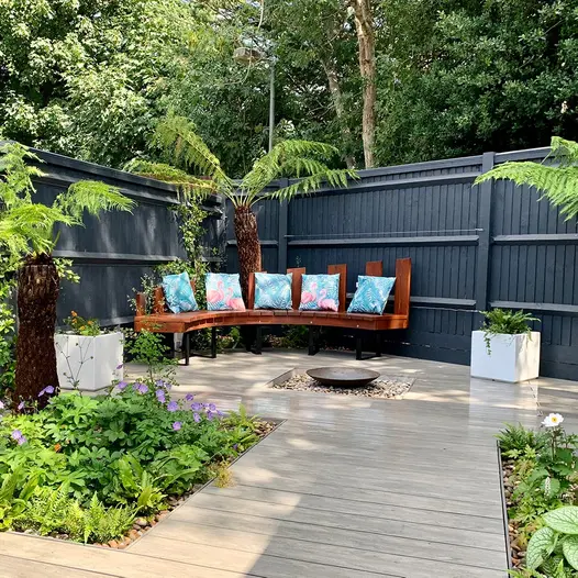 Curved wooden bench sits against black fence with luna designboard composite decking on the floor with a water feature in the middle.
