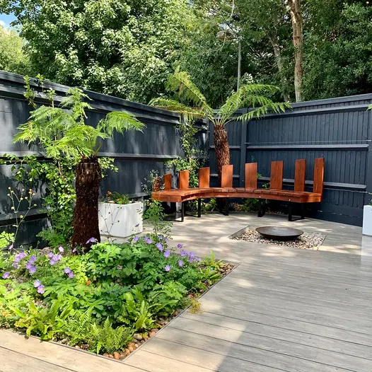 A curved wooden bench rests against a black fence, with Luna designboard composite decking underfoot and a water feature in the center.