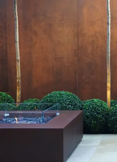 Two panels of steel Corten DesignClad sees young tree stumps with topiary bushes and firepit below.