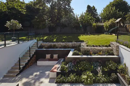 A modern garden in a sunken courtyard with three tiers, bullnosed porcelain steps, and retaining walls, with plants in the background.