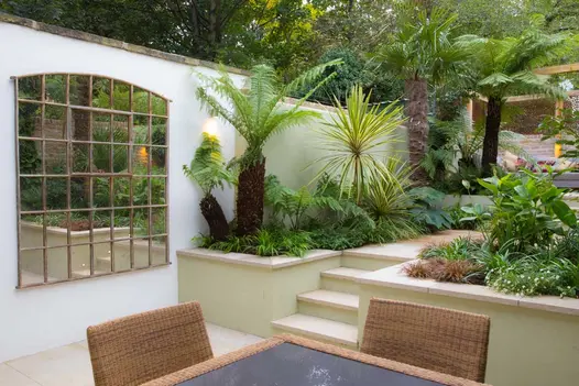 Large mirror emulates window on white wall of patio. To right, 4 steps rise between exotic planting, leading towards pergola.