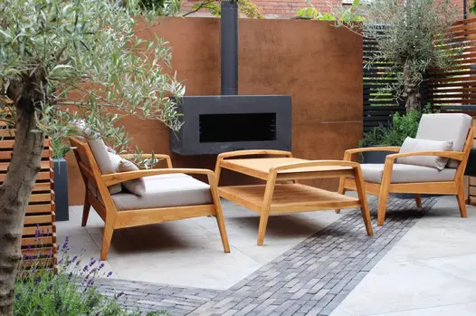 4 steel corten DesignClad panels clad large wall with built in chimenea with 2 armchairs and a table next to it.