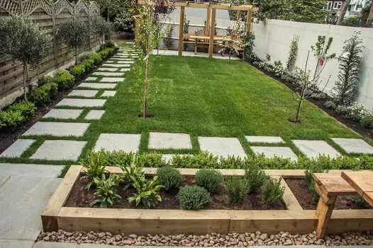 Oblong lawn with Kandla Grey sandstone slabs laid as stepping stone on 2 sides. Matching patio at far end with wooden pergola.