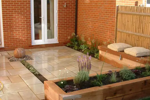 Patio of Harvest sawn sandstone outside french doors, with water feature set on pebbles and earth rill running towards raised bed.