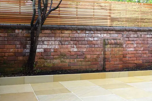 A back garden with Harvest Edging Stones, matching sandstone paving, and a leafless tree against a brick wall.