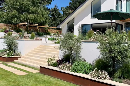 10 Harvest sawn sandstone steps rise between flanking walls from lawn to patio of white-walled house with slant-top windows.