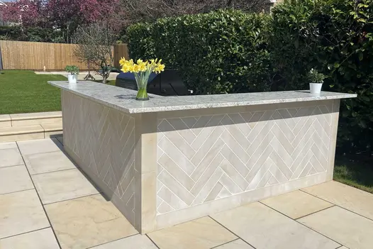 Vase of daffodils sits on right-angled outdoor bar by hedge, on Harvest Smooth sandstone paving, with 2 steps up to lawn.