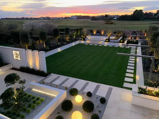 Modern, rectilinear garden with square lawn surrounded by white-walled raised beds and areas of Harvest Sawn Sandstone paving.