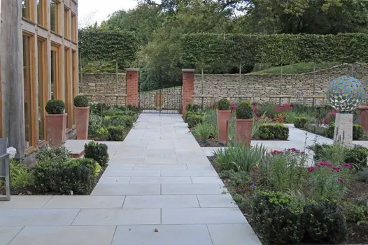 Wide path of Harvest sawn sandstone paving leads past building to gate between 2 brick pillars in dry stone wall. 