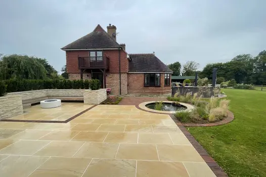 Contemporary patio featuring Harvest Sawn Sandstone with a warm, golden hue, accented by brick edging, leading to a traditional brick house with a landscaped garden.