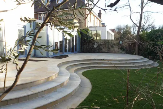 Large, bespoke circular patio in the shadows showing of harvest smooth sandstone pavings multiple tones of creams and beiges.