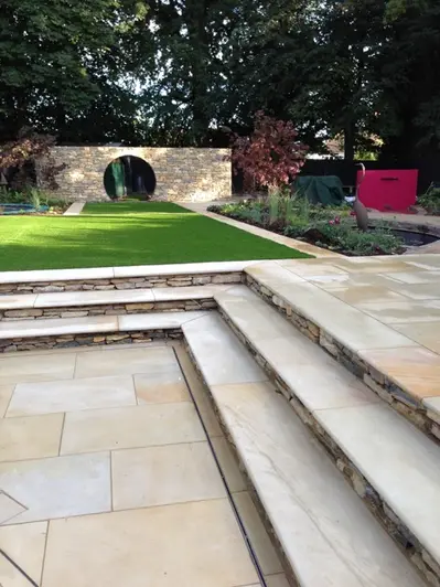 Corner of patio with 3 steps rising on 2 sides to Harvest sawn sandstone paving, with lawn and moon gate in background.