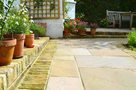 Low view across Autumn Brown Indian sandstone paving slabs edged on 2 sides by 2 steps of bricks with planted pots on them. 
