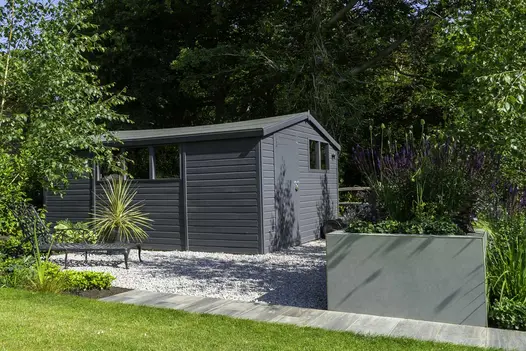 Tall planting trough faced with Hydra Plomo DesignClad sits in shrub-planted bed by gravel area with grey garden building.