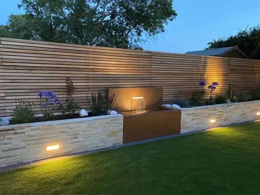 dusk views show water feature in between sandstone clad raised planter walls, clad with steel corten designclad.