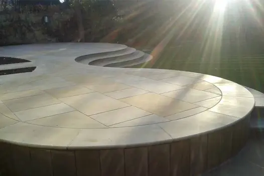 Large, bespoke circular patio glistens in the sunlight showing of harvest smooth sandstone pavings multiple tones of creams and beiges.