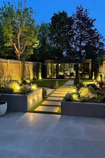 Shot at night, view down a garden shows planks of jura beige sawn limestone leading from patio to garden building.