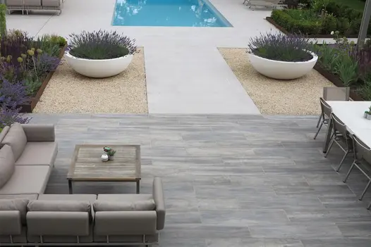 Garden dining and lounging furniture on a porcelain wood effect patio with a pathway leading towards a swimming pool.