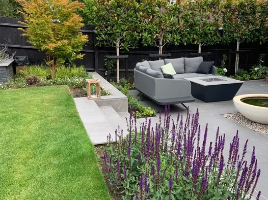 Raised lawn area surrounded by flower beds  and looking down onto a Steel Grey porcelain patio and lounging furniture.