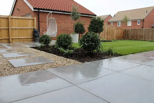 White swing chair hangs over Anthracite 800x800 porcelain paving, next to lawn, bed with clipped bushes, and slabs set in gravel.