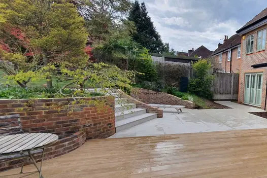 Decking space adjacent to Area 800x800 Porcelain Paving used with steps leading up to lawn. Large Paving slabs with red brick wall.