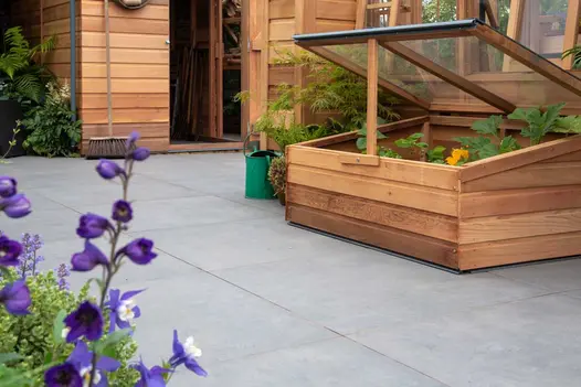 A broom leans against wooden shed, next to greenhouse. Cold frame sits on Anthracite Large Patio Slabs. Design by Base Squared.