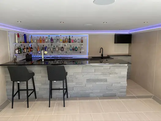 Black barstools sit at stone clad bar, wall of drinks with sink and speakers using desert beige composite cladding.