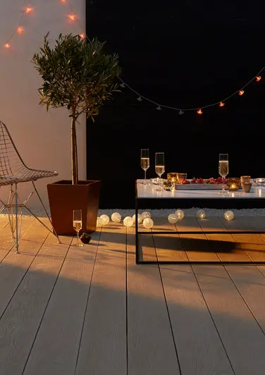 Champagne glasses and string lights adorn low table with metal chair sat on limed oak millboard decking with corten planter.