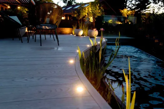 Night view shows smoked oak millboard curve around a pond with built in flowerbeds, decking has spotlights lining the edge of the pond.