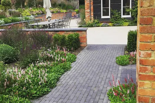 Path of Lucca Dutch clay pavers runs past planted border to L-shaped white patio wrapped around corner of house.