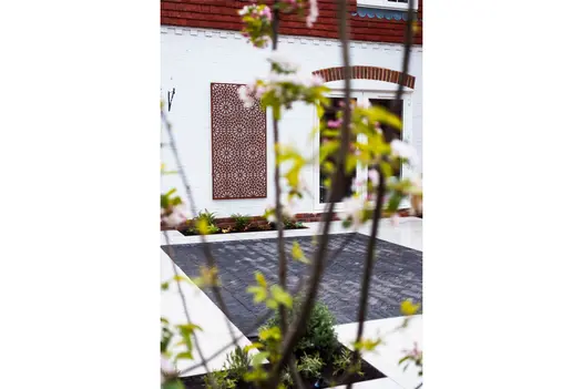 View through blurred flowering twigs to pale patio with square of grey Lucca clay pavers opposite a wall hanging.